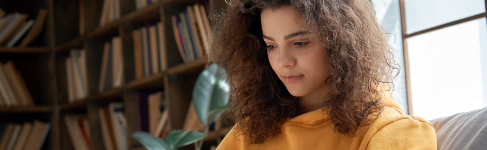 Woman in a yellow sweater and blue jeans holding a mug and reading a laptop while sitting on a grey couch next to a cat.