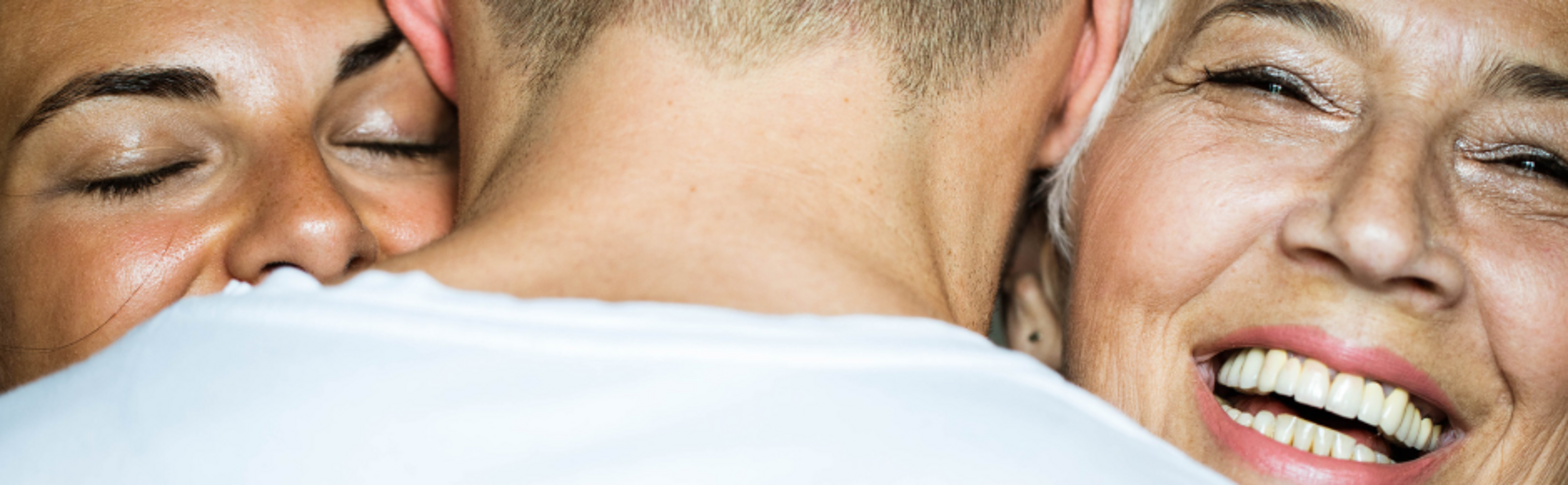 Man in white shirt being hugged by a woman with brown hair and another woman with white hair.