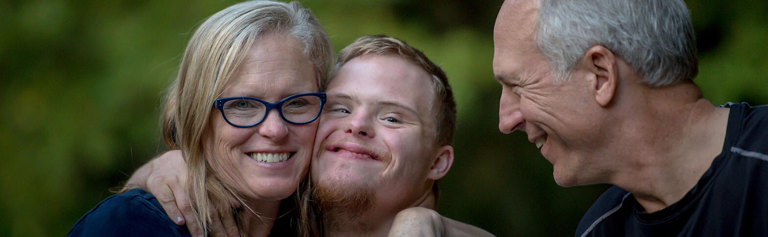 Family of three pose for a photo together. Mother and son have their arms around each other and faces pressed against each other. The father is in profile view looking at both of them smiling.