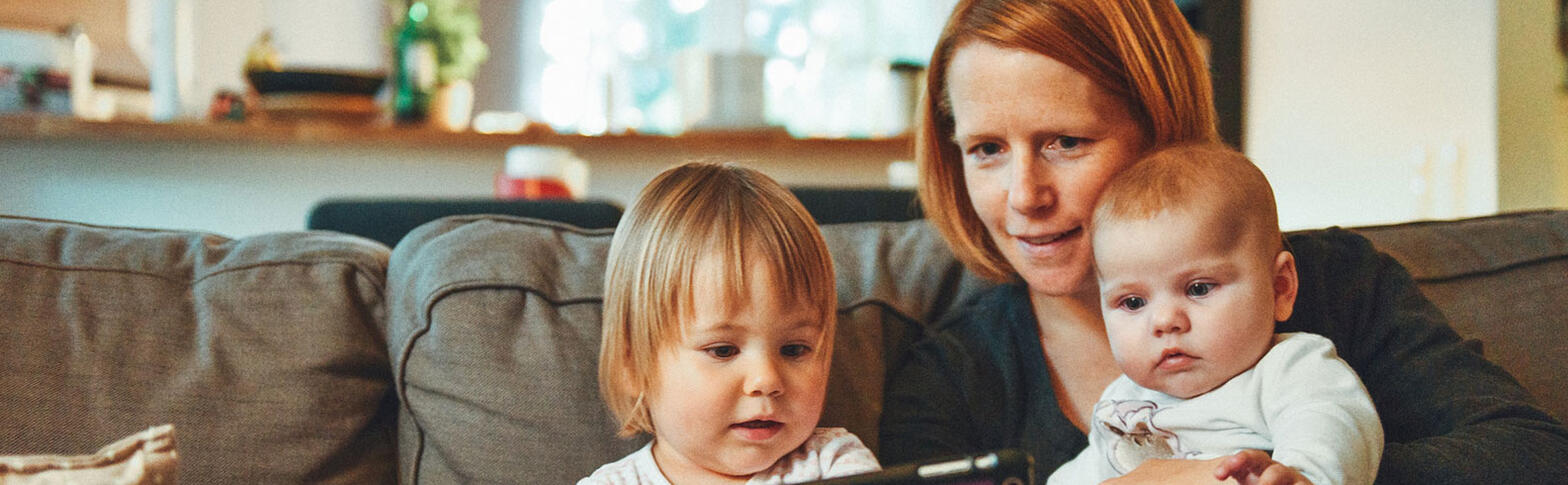 Two children, one a baby and one a toddler, sit on their mothers lap. Together, on the couch, they are looking at something on a tablet.