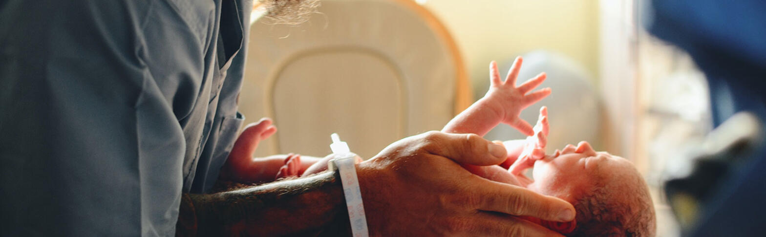 New born baby in a hospital, who is in a man's hands, reaches up and spreads their fingers wide.