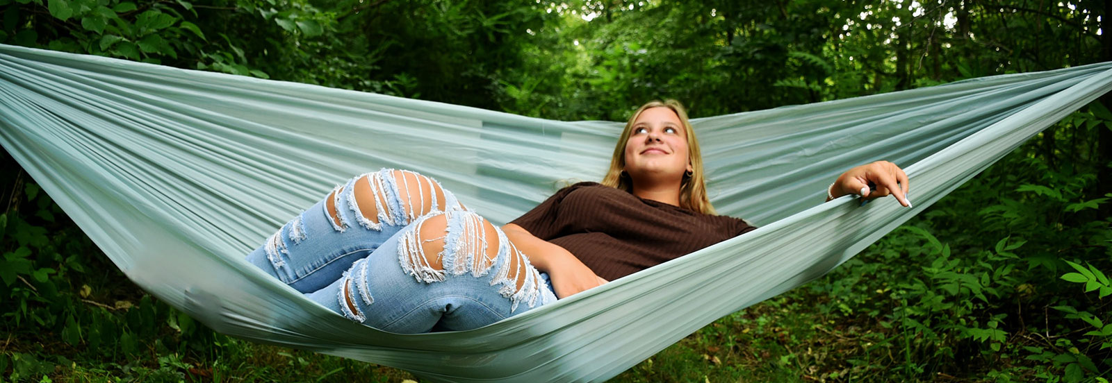 Teenage girl laying in a teal blue hammock. Green grass and foliage surrounds the hammock.