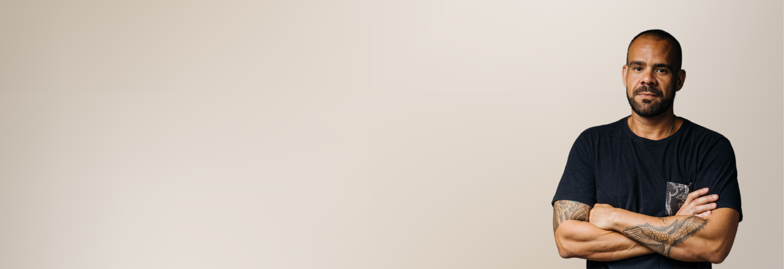 A man with short buzzed hair, close beard and tattoos with crossed arms and a serious expression. 