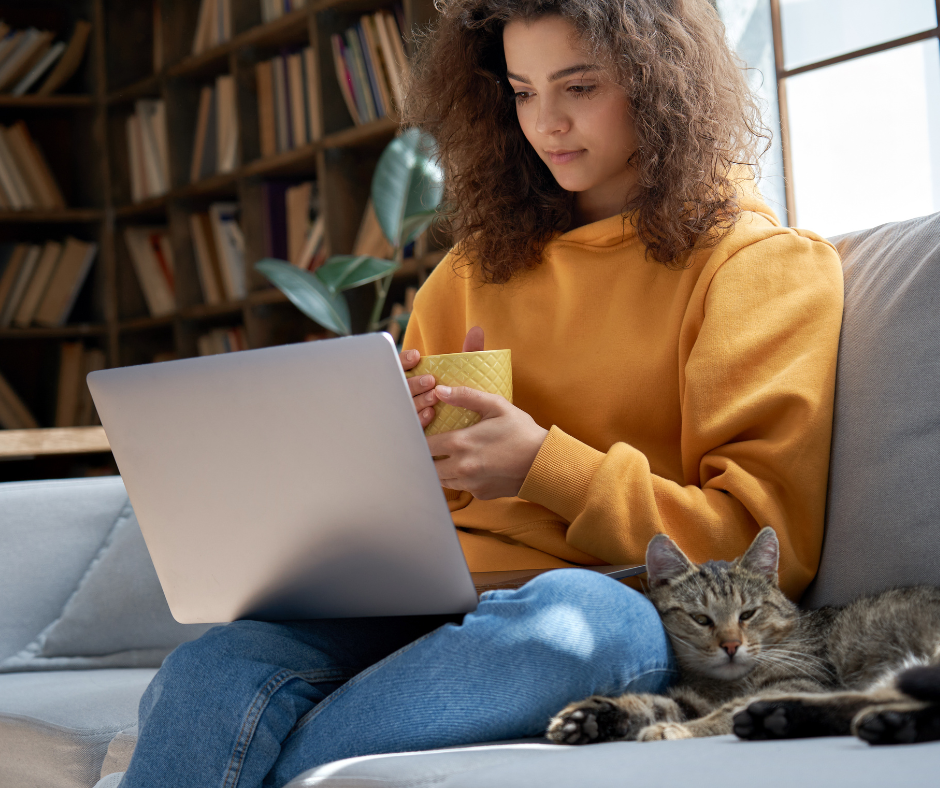 Woman in a yellow sweater and blue jeans holding a mug and reading a laptop while sitting on a grey couch next to a cat.