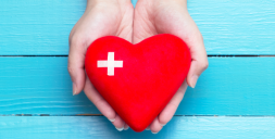 Hands holding a red heart-shaped object with a white medical cross on it.